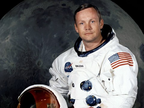 Portrait of Astronaut Neil A. Armstrong, commander of the Apollo 11 Lunar Landing mission in his space suit, with his helmet on the table in front of him. Behind him is a large photograph of the lunar surface. (Image Credit: NASA)